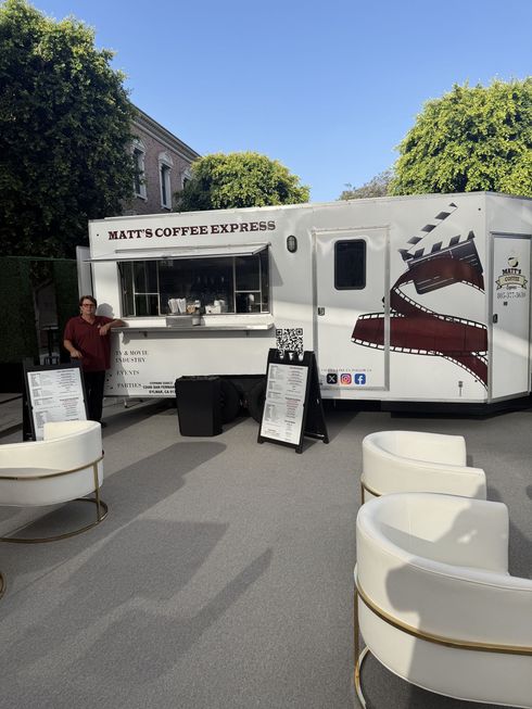 Outdoor food truck with coffee and drinks, white seating in front, and a person serving at the window