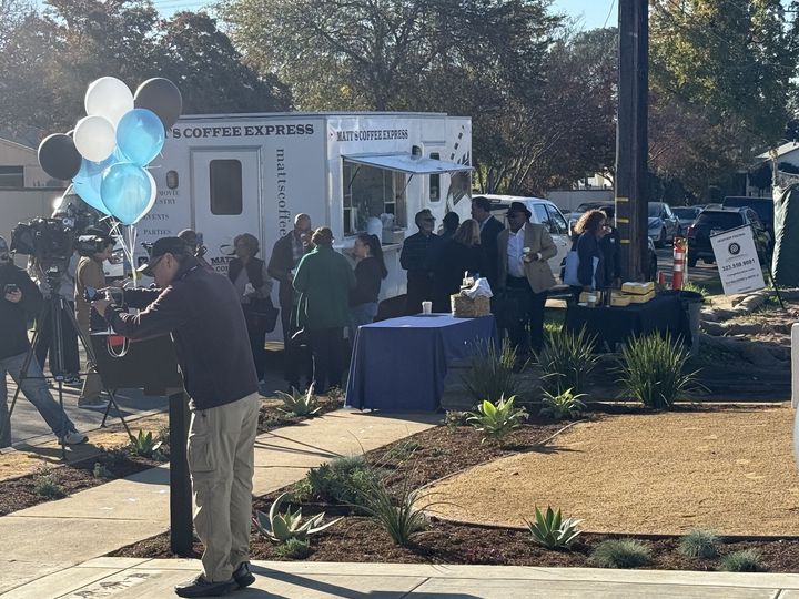 Outdoor community event with people gathered around white tents and blue balloons on a sunny day