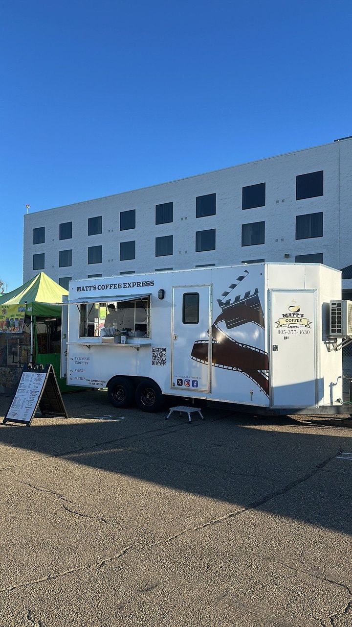 White food truck beside a multistory building on a gravel lot, with a green canopy nearby.