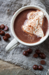 White mug of hot cocoa topped with whipped cream and cinnamon on a rustic table with coffee beans