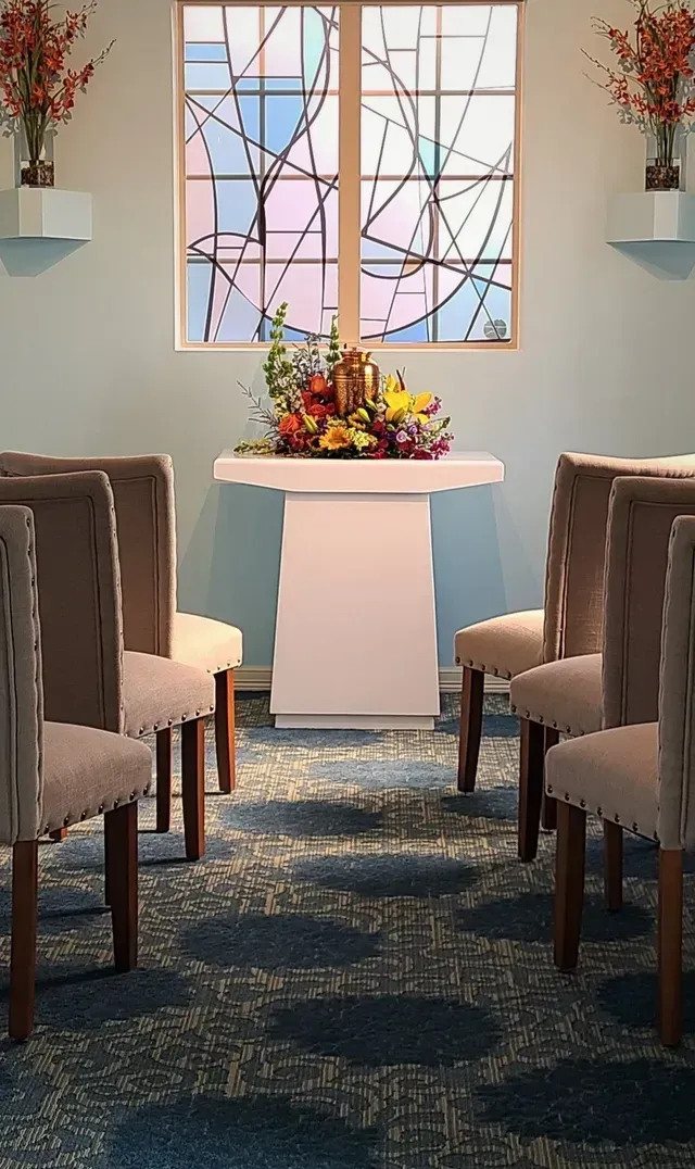 Symmetrical chapel aisle with upholstered chairs, a small altar table, and a window behind floral arrangement