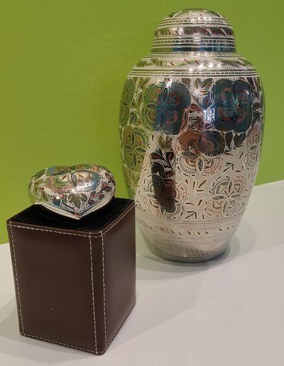 Two silver engraved urns on a white surface, one large and one small on a brown stand, against a green background