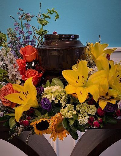 Colorful floral arrangement with yellow lilies, orange roses, sunflowers, and a dark urn on a table