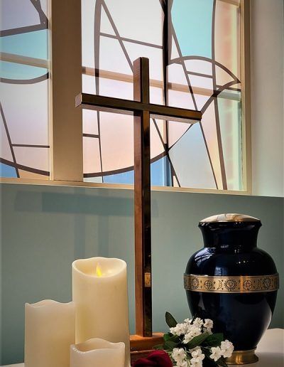 Church altar with cross, candles, white flowers, and a dark blue urn in front of stained glass.