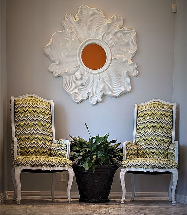 Two patterned chairs flank a potted plant beneath a large white decorative wall mirror.