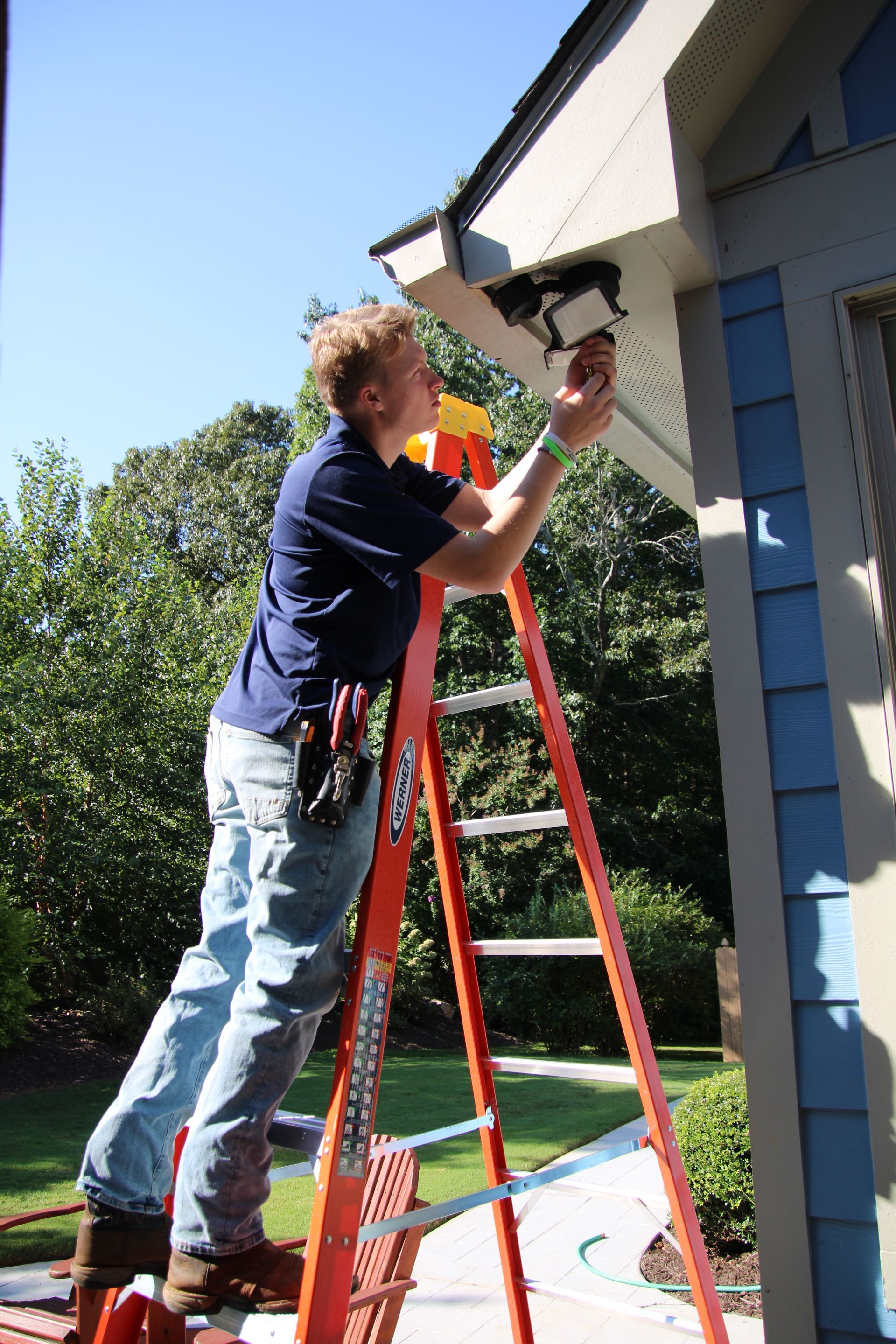 Person on a red ladder using a power drill to work on the side of a house outdoors.