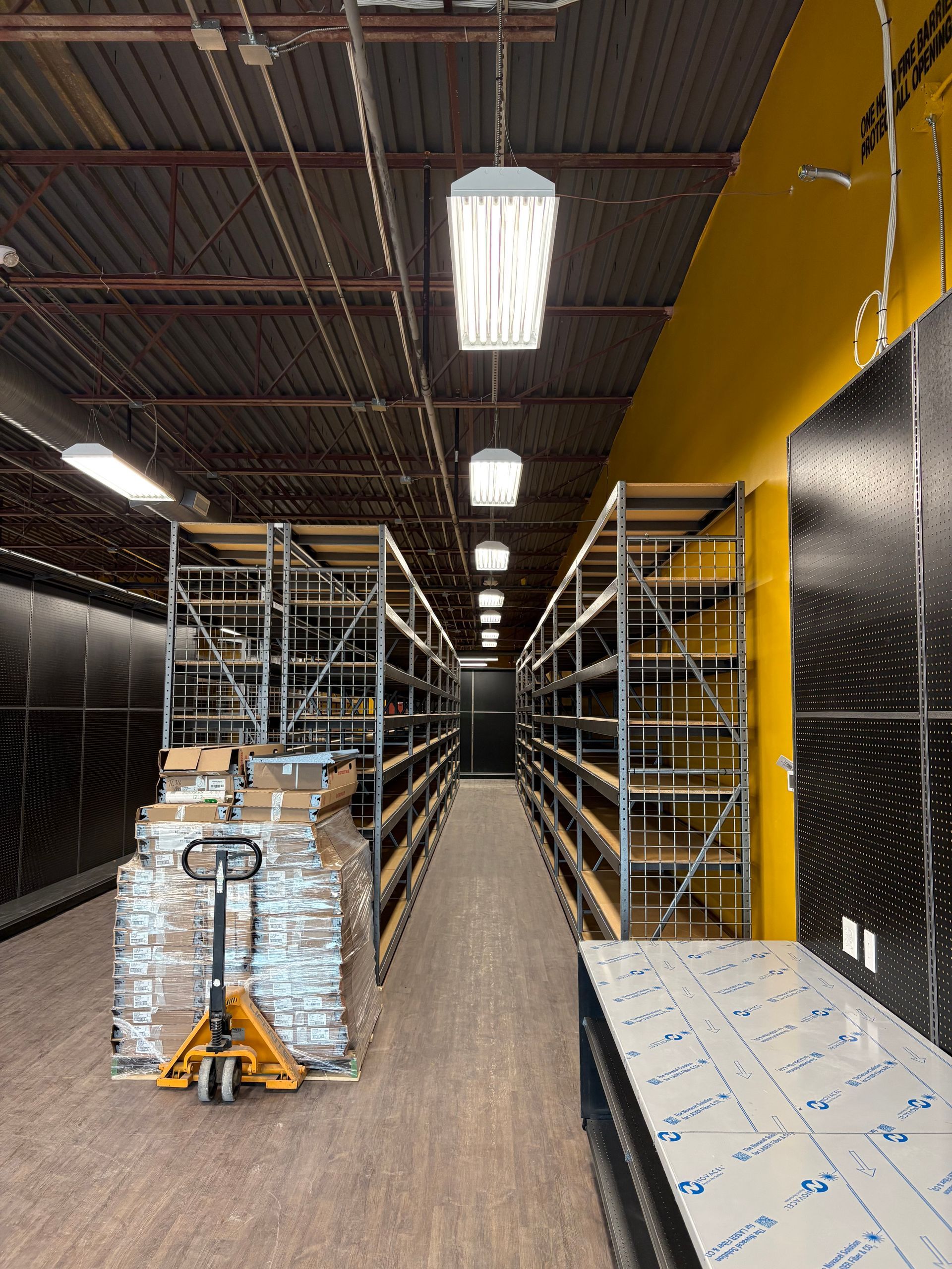 Warehouse aisle with tall metal shelving, pallet jack, and boxes under bright overhead lights.