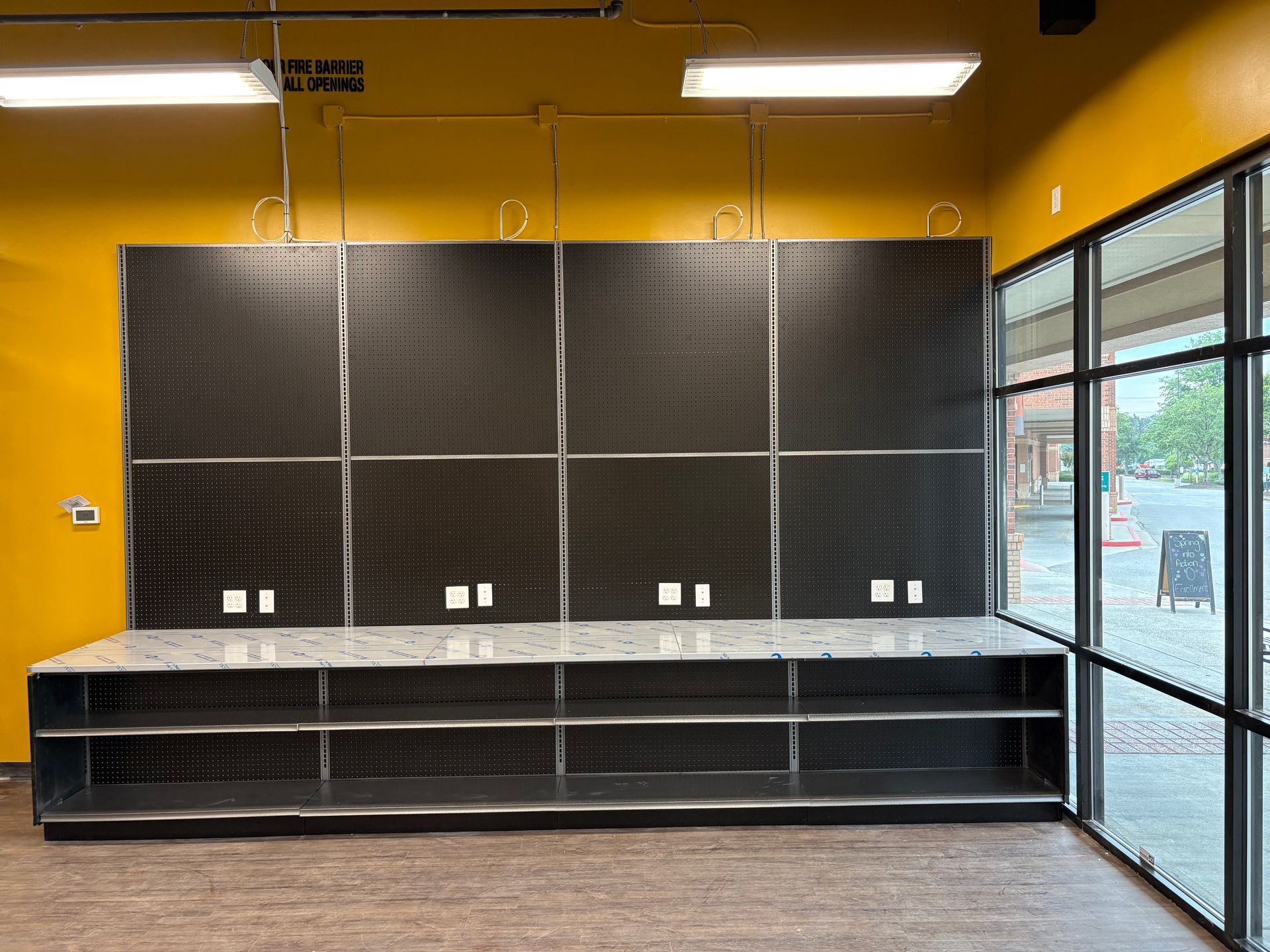 Empty retail checkout counter with black pegboard wall, yellow walls, and large front windows