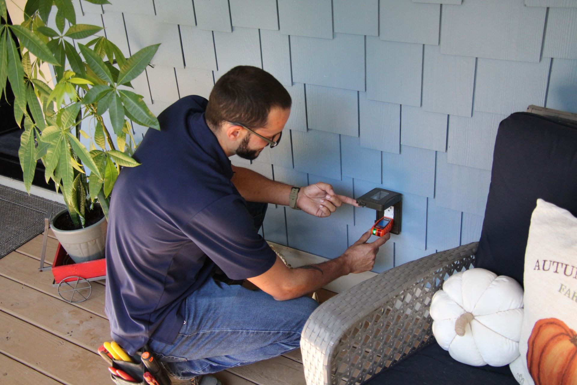 Man kneeling to plug a device into an outdoor wall outlet beside a patio chair and potted plant