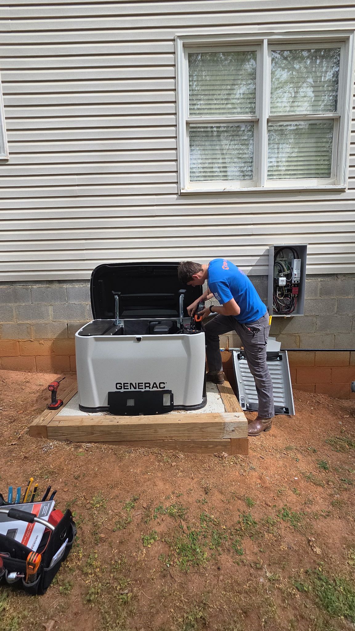 Person working on a generator beside a house exterior, with tools on the ground nearby.