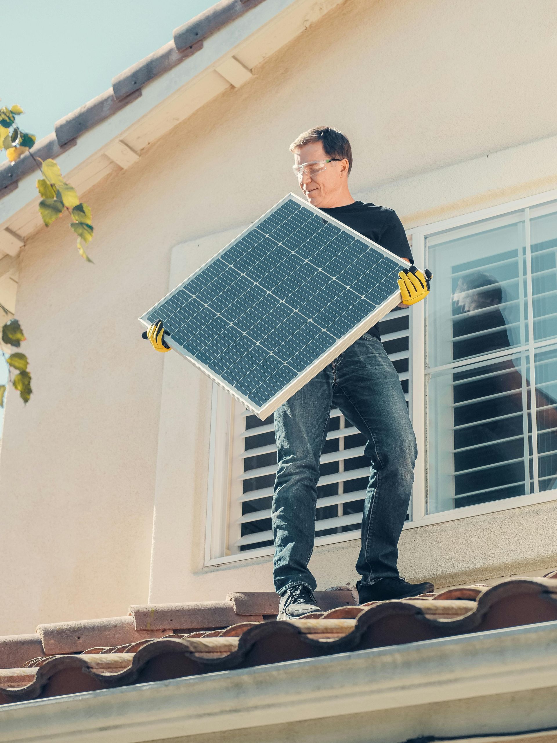 Man installing a solar panel on a rooftop, wearing work gloves, in front of a window. Sunny day.