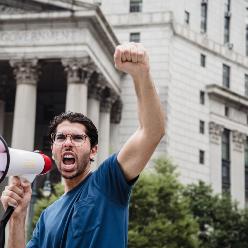 Man with megaphone raises fist, shouting in front of a government building.
