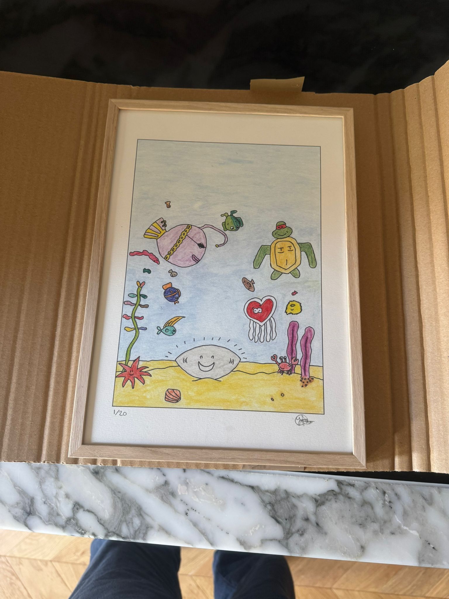 Kleurrijke kindertekening van zeeschildpadden en zeeleven in een onderwateromgeving, ingelijst tegen een beige muur.