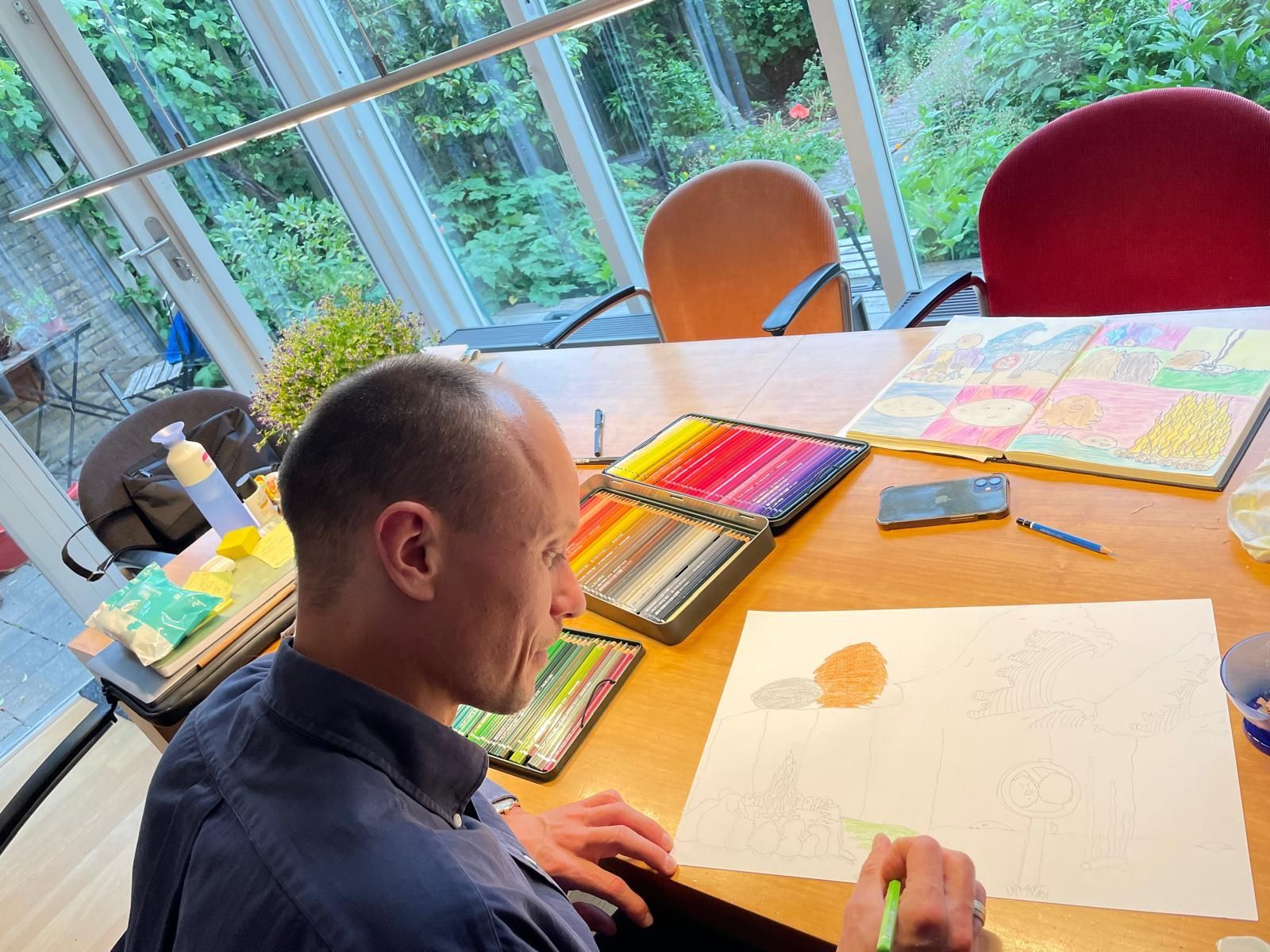Een man tekent aan een tafel met tekenmateriaal. Heldere kleuren, natuurlijk licht en uitzicht op groen door het raam.