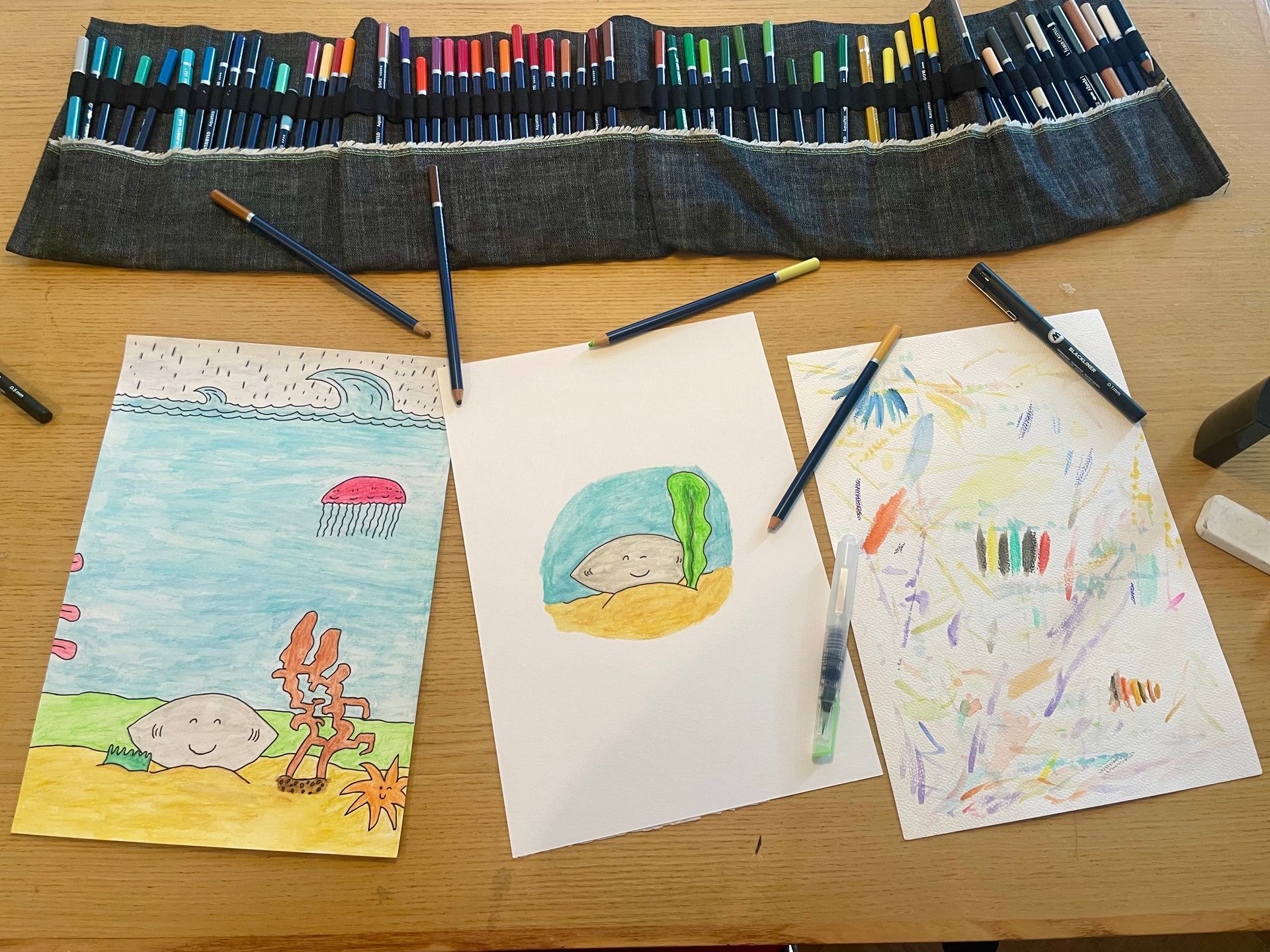 Drie kleurrijke tekeningen met kleurpotloden liggen op een tafel. Boven de tekeningen staat een etui gevuld met potloden.