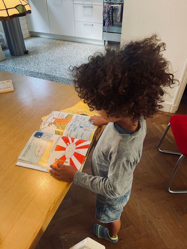 Kind met krullend haar leest een kleurrijk boek aan een tafel.