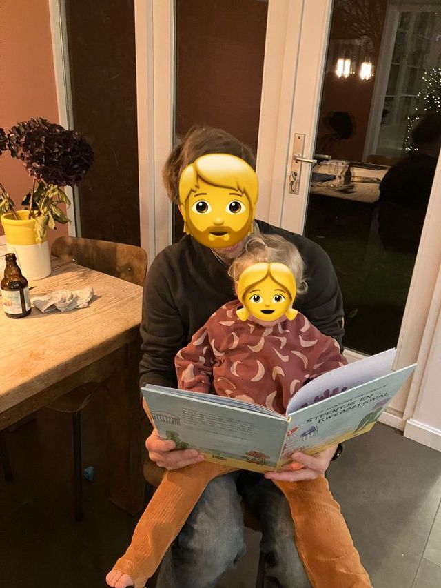 Een persoon houdt een kind vast, beiden kijken naar een open boek. Binnen, warm licht.