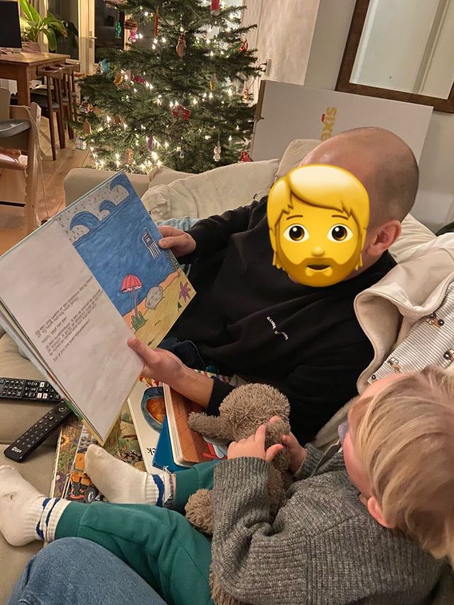 Iemand leest hardop voor uit een boek aan een kind, zittend op een bank met een kerstboom op de achtergrond.