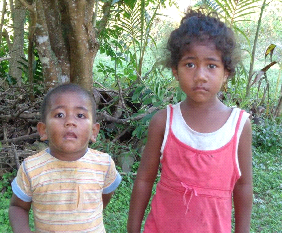 Two children stand outdoors, one in a striped shirt, the other in a red dress. They both look towards the camera.