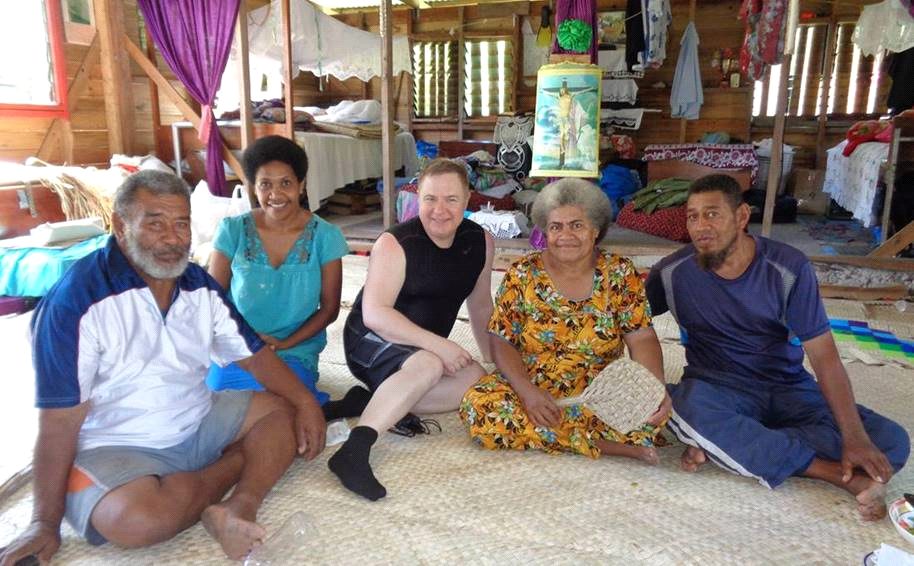 Five people seated on woven mat inside a wooden structure; smiling, with a variety of clothing and skin tones.