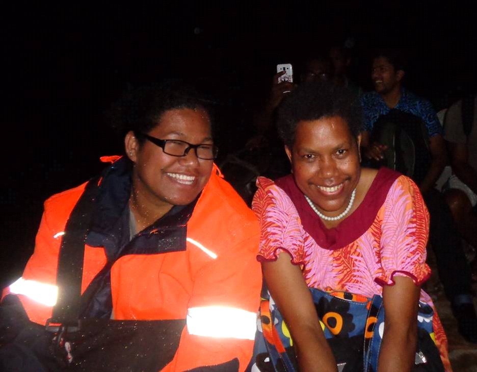 Two smiling people sit close together outdoors at night. One wears orange jacket, the other a pink patterned dress.