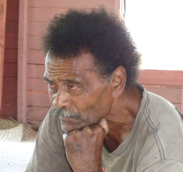 Man with dark skin, holding chin, looking down thoughtfully; indoor setting with wooden walls.