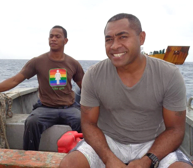 Two men on a small boat at sea. One smiles; the other looks forward. A rusty ship is in the background.