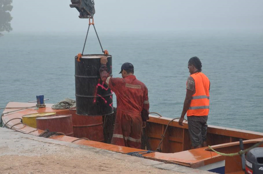 Workers loading a black barrel onto an orange boat under a crane near a body of water in the rain.