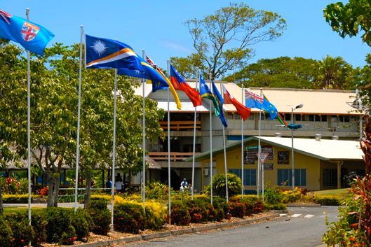 Flags of various nations fly in front of a building with a yellow facade and a green lawn.