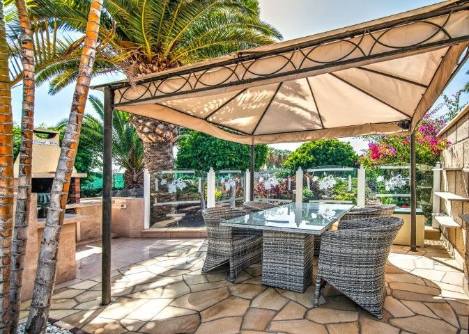 Outdoor island eating area with wicker chairs and a tan colored sail sun cover to block sun. set in backyard with palm trees