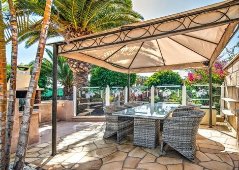 Outdoor island eating area with wicker chairs and a tan colored sail sun cover to block sun. set in backyard with palm trees