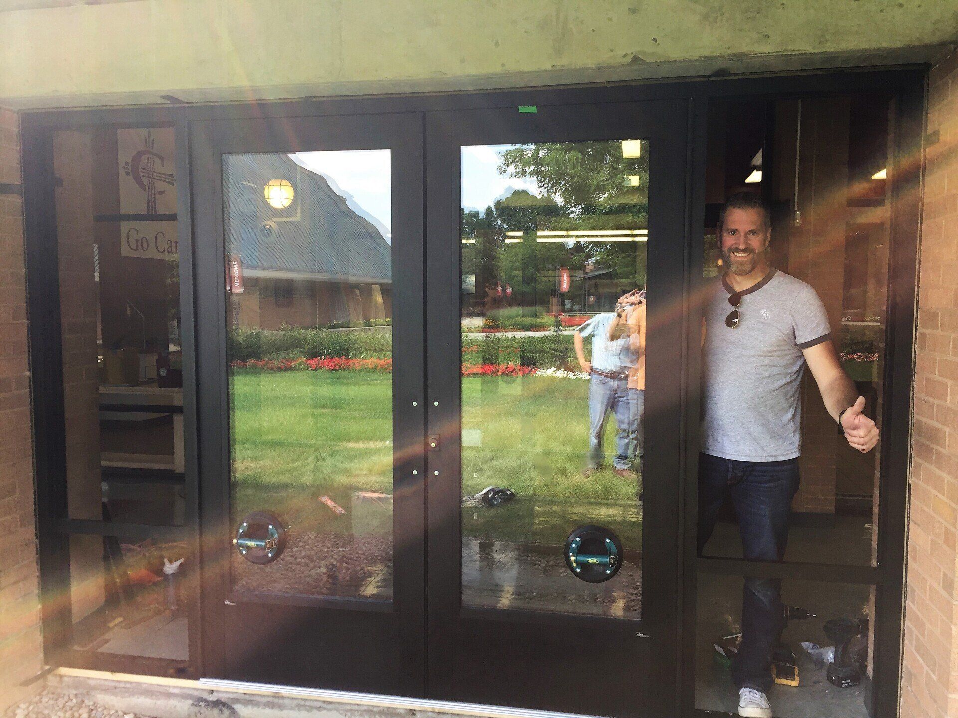 Man standing in doorway, smiling. New black double doors with glass panels reflecting a green lawn.