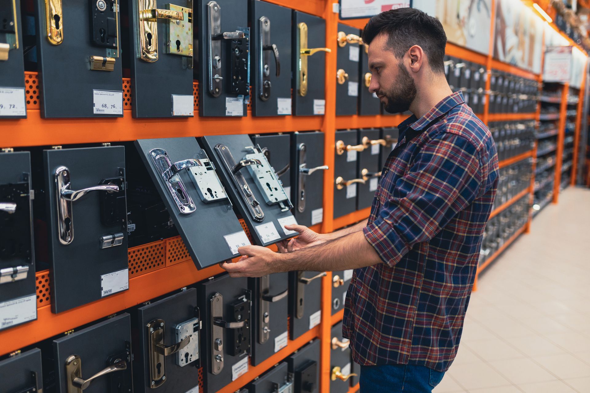 Construction shop shopper next to rack with doorknobs and locks. Man in plaid shirt examines door locks in a hardware store aisle.