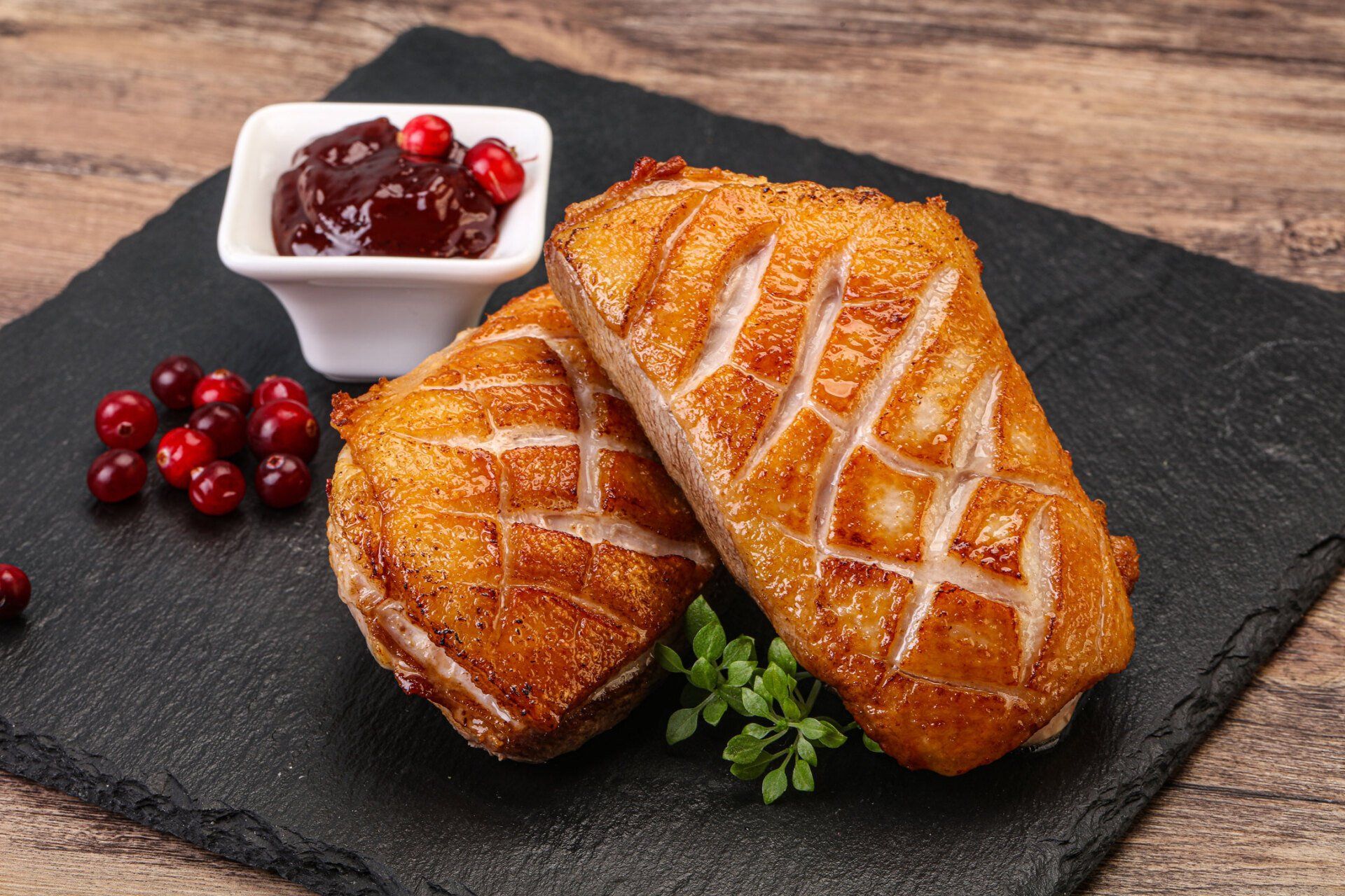 Twee krokant gebakken eendenborstfilets met cranberrysaus en verse bessen op een zwarte leistenen plank.