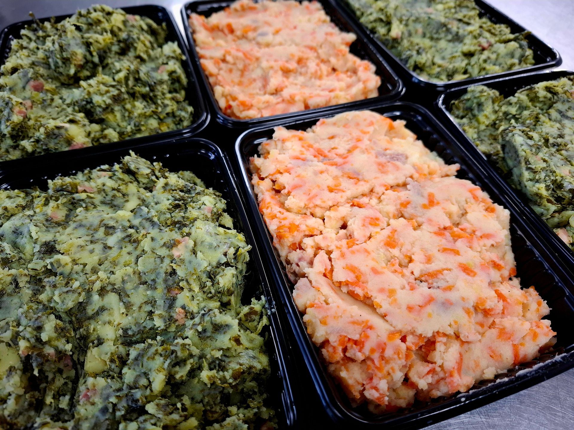 Zwarte bakjes gevuld met gepureerde groenten, waaronder oranje wortelpuree en groene boerenkool- of spinaziepuree.