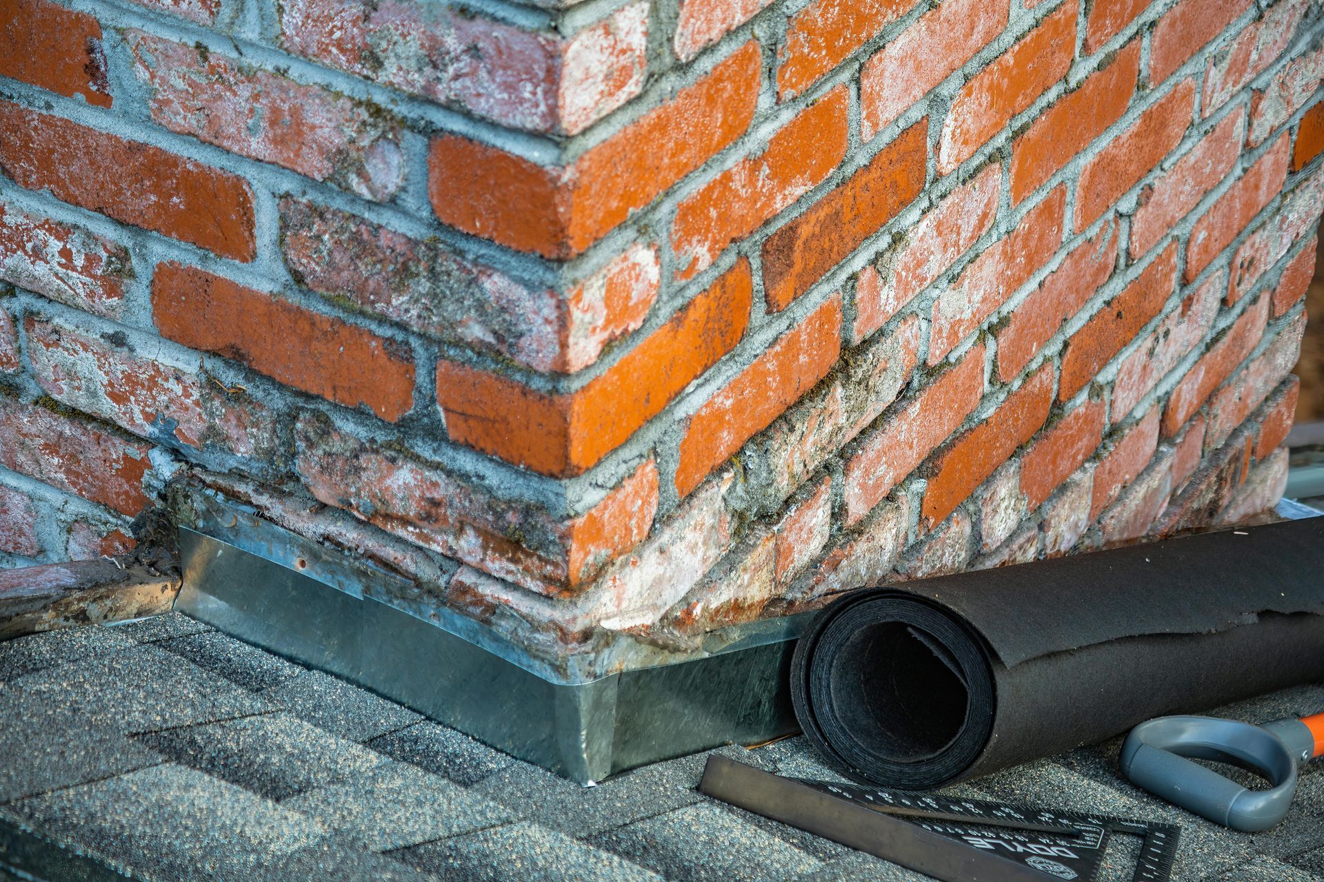 Brick chimney base on a rooftop with black pipe and gray flashing repair material