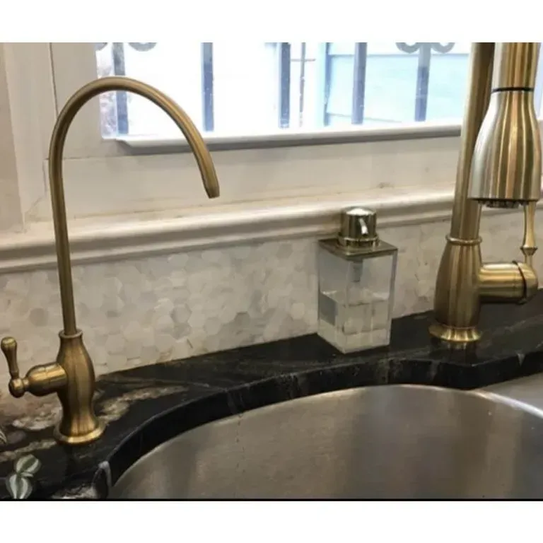 Kitchen sink with two brass faucets and a small soap dispenser on a black countertop by a window