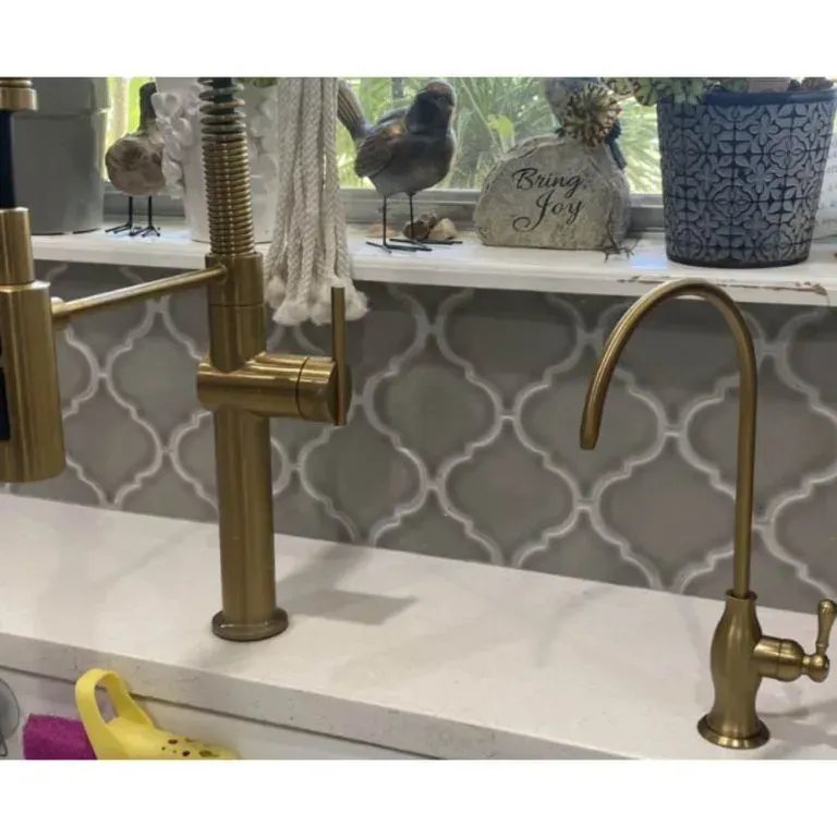Gold faucet and soap dispenser on a white kitchen sink with patterned tile backsplash and window decor