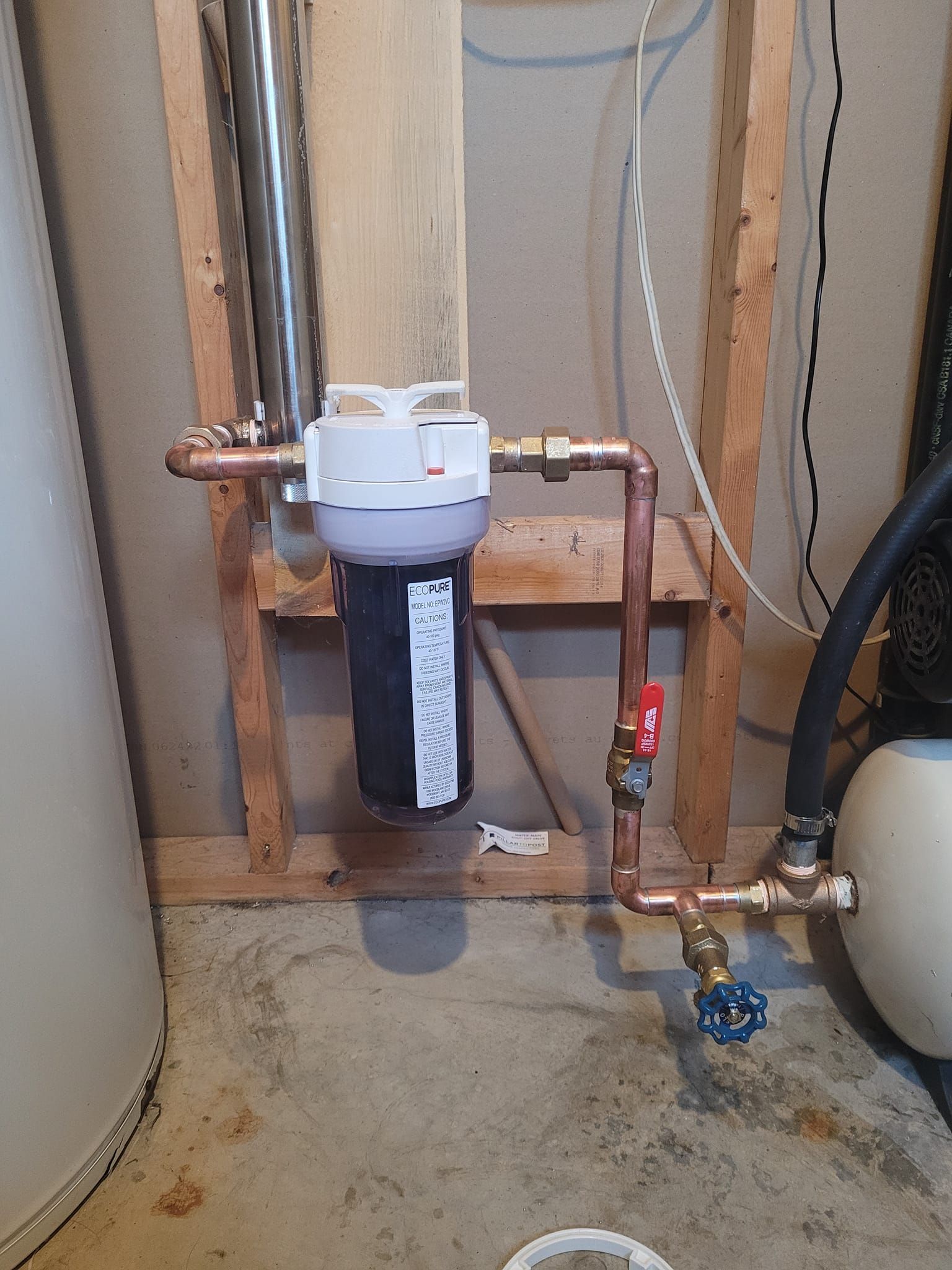 Water filter system with copper pipes, a red valve, and a blue valve. Located near a water tank on a wood-framed wall.