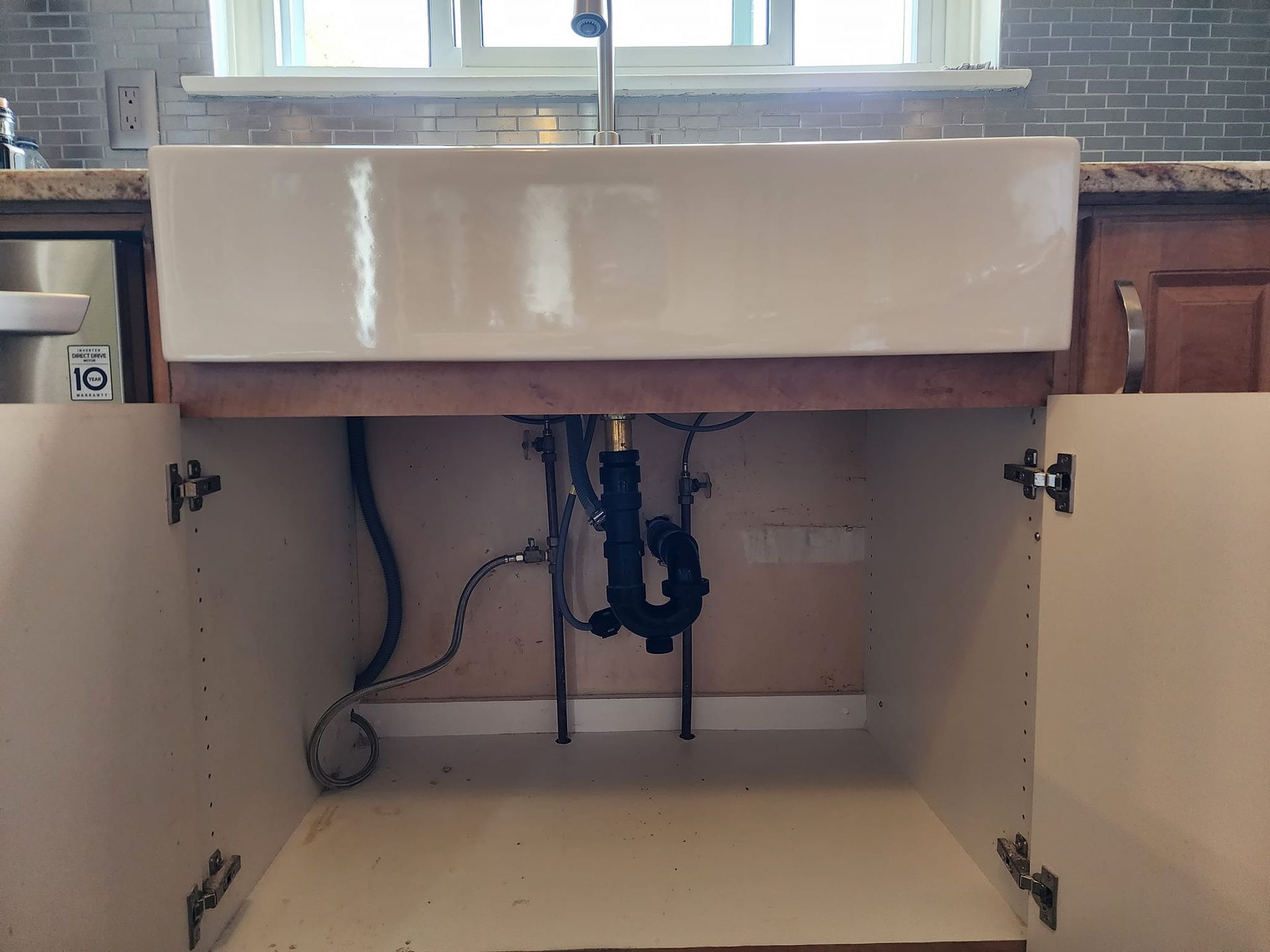 Open kitchen cabinet under a farmhouse sink, showing plumbing and support structure. Beige, wood, and black.