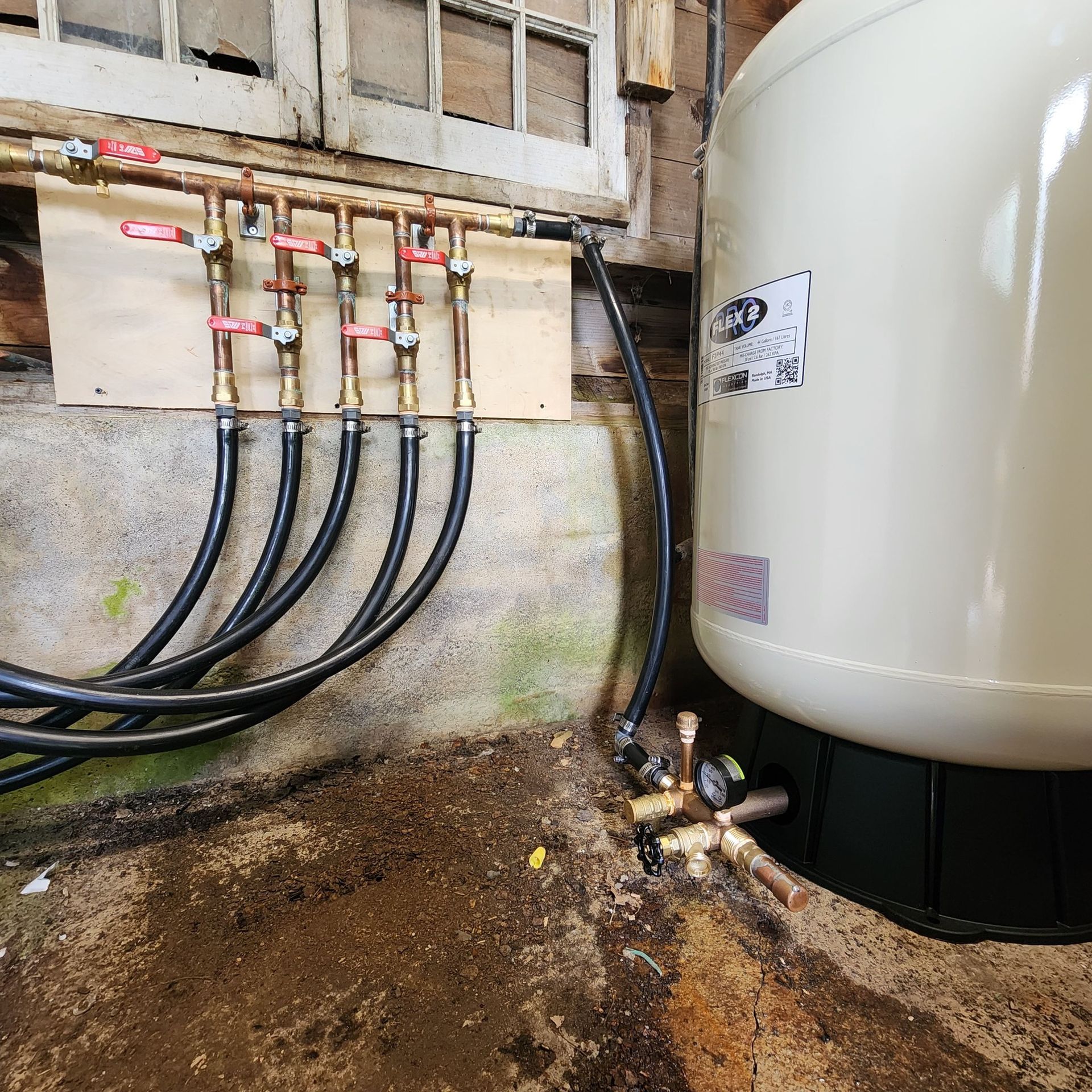 Plumbing system with copper pipes, black hoses, and a white pressure tank in a damp basement.