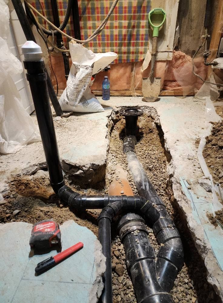 Black plumbing pipes in a trench, likely for a bathroom. A tape measure and marker lie nearby.