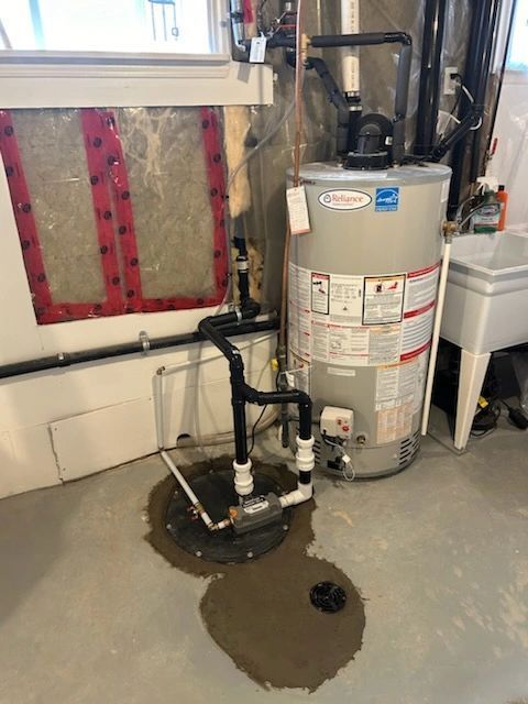 Water heater and sump pump in a basement with wet concrete floor and exposed insulation on the wall.
