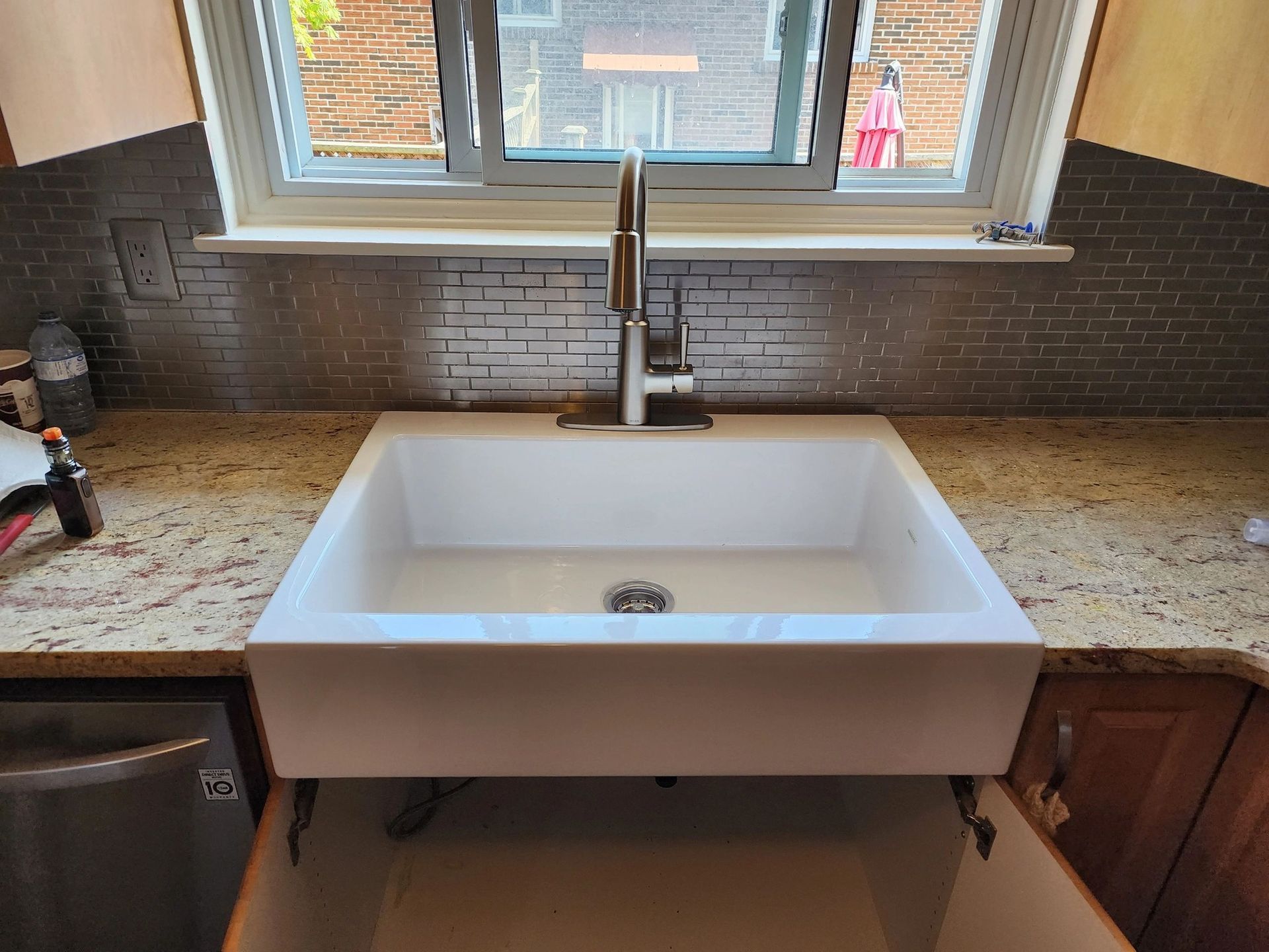 White farmhouse sink with faucet in a kitchen, beneath a window with granite countertop, cabinet doors open.