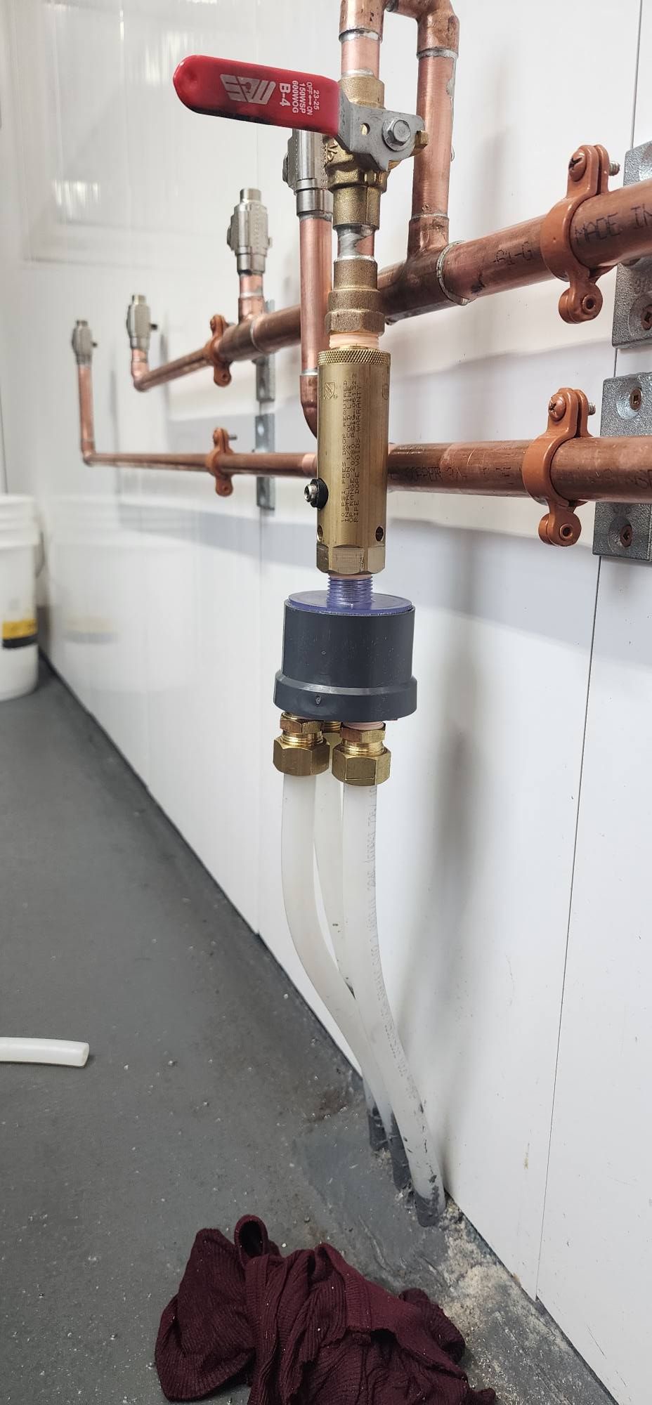 Copper pipes with valves and plastic tubes attached to a wall. A red valve handle is visible.