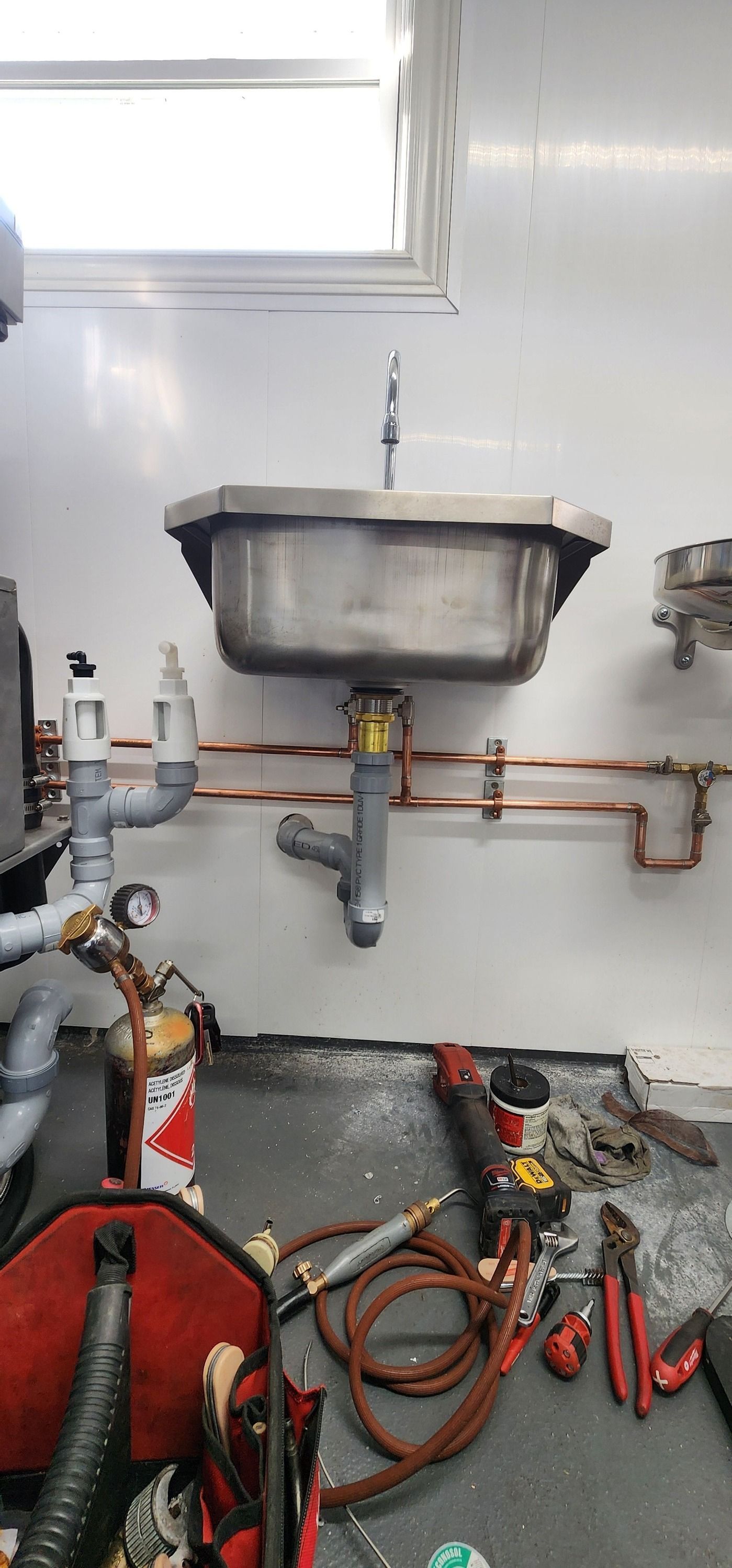 Stainless steel sink with plumbing, tools, and a red tool bag, likely in a workshop.