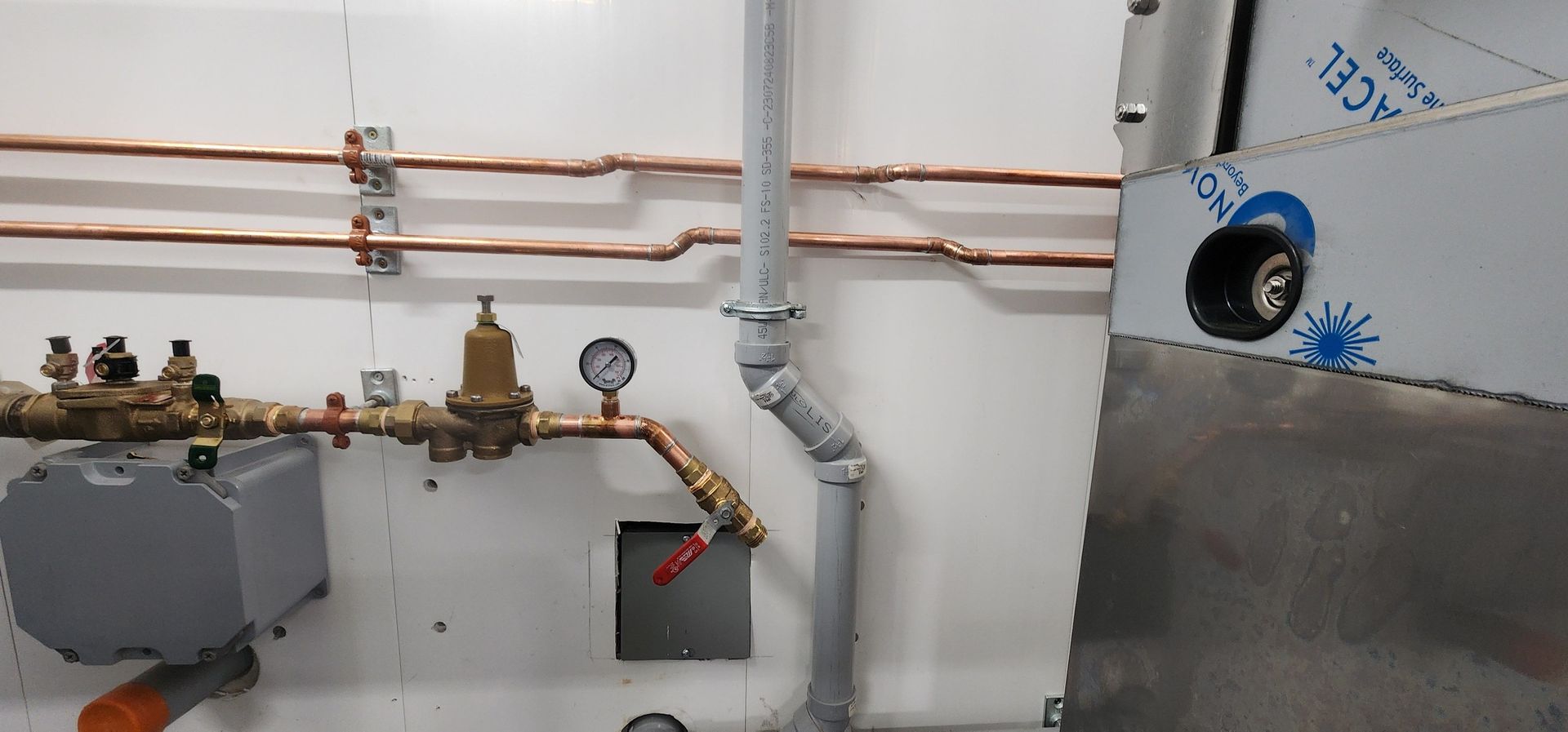 Piping and valves on a white wall; gray conduit, copper pipes, gauges.