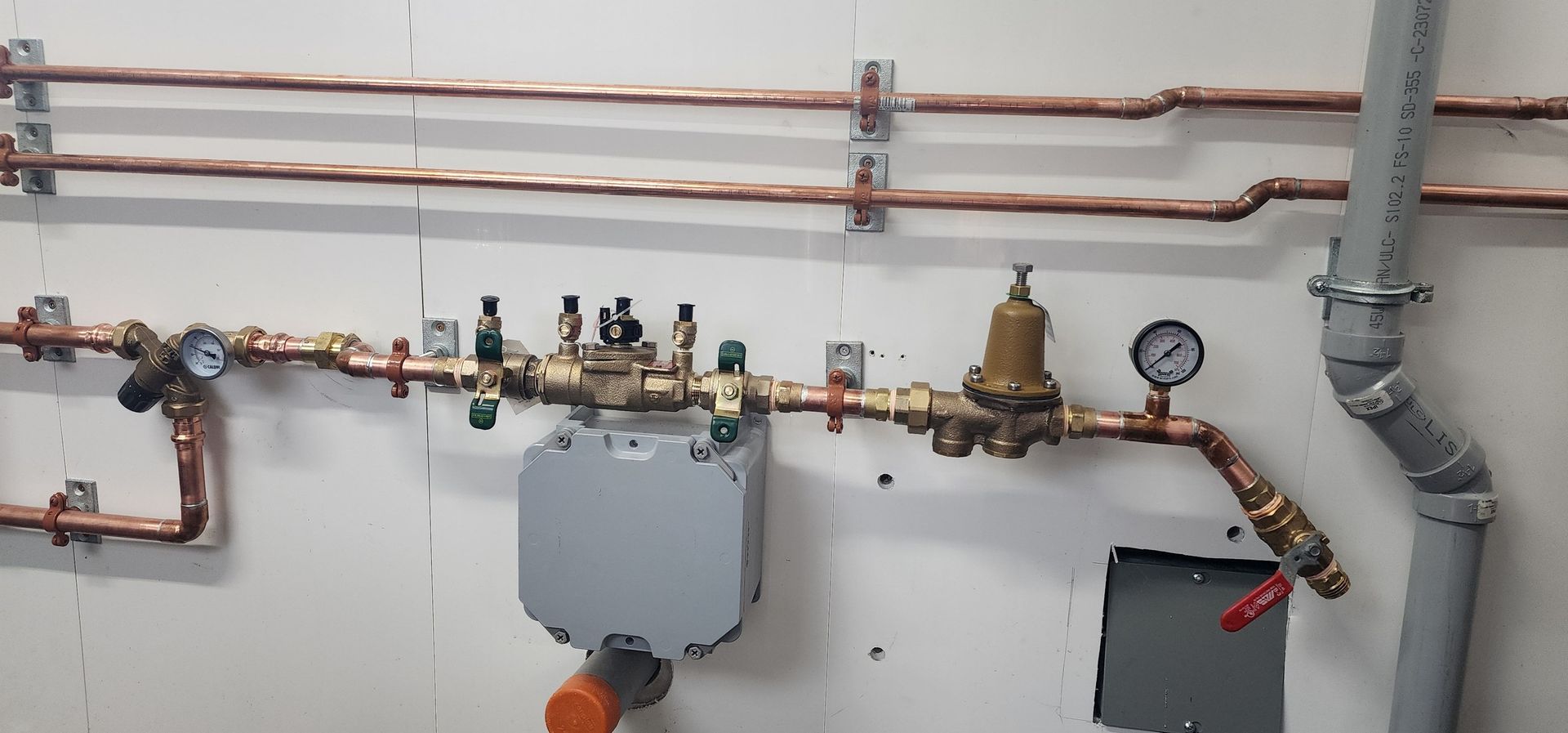 Copper pipes and plumbing fixtures against a white wall. Includes a pressure gauge and other components.