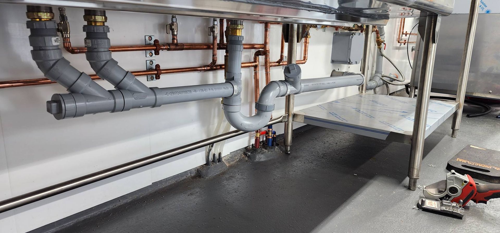 Plumbing under a stainless steel sink with copper pipes and gray drain pipes.