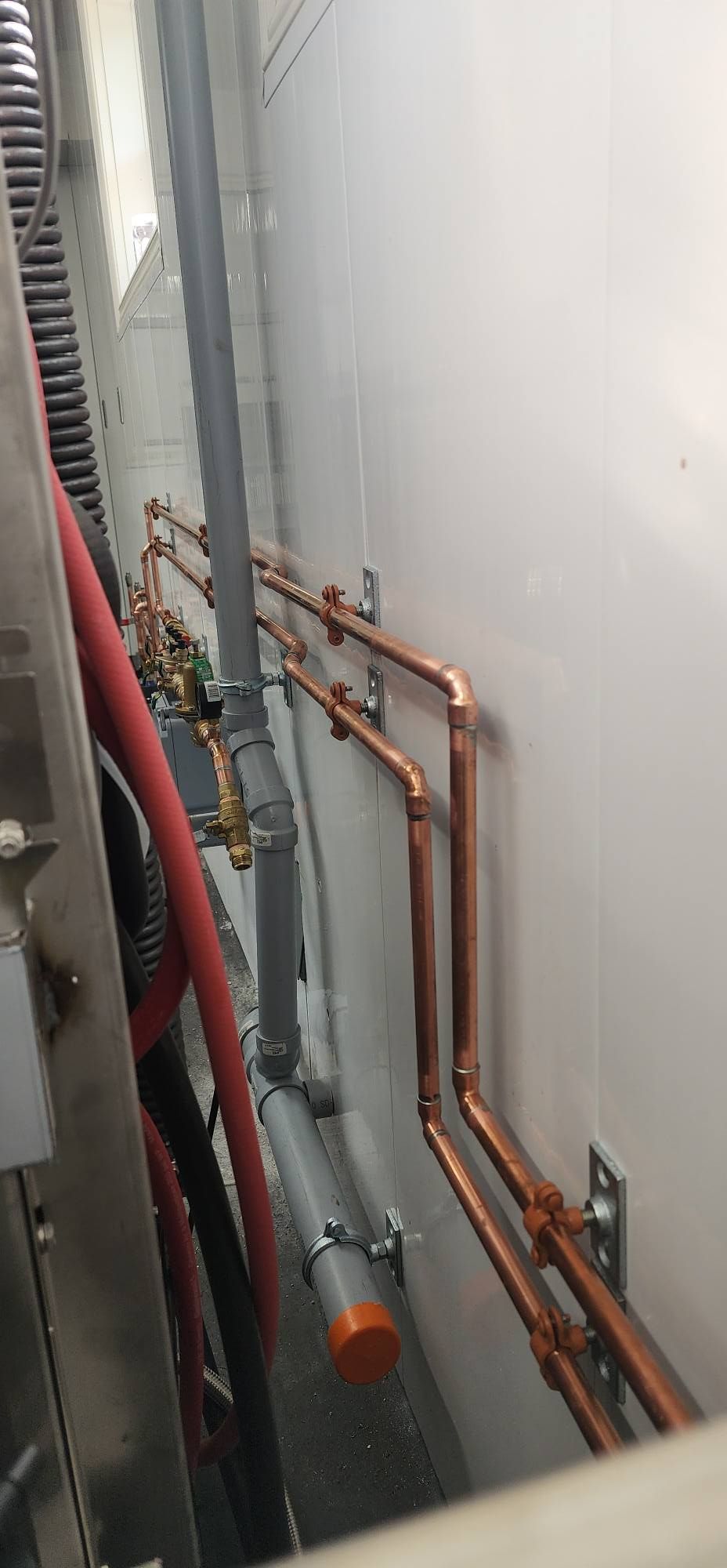 Copper pipes along a white wall, with gray and red pipes next to them.